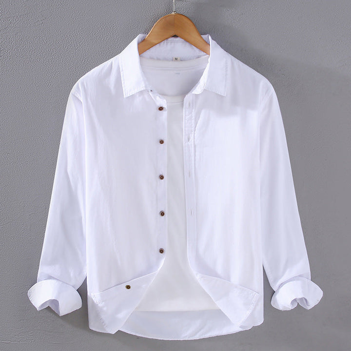Camicia bianca da uomo in cotone, maniche lunghe, colletto classico, bottoni marroni, appesa su gruccia di legno, sfondo grigio.