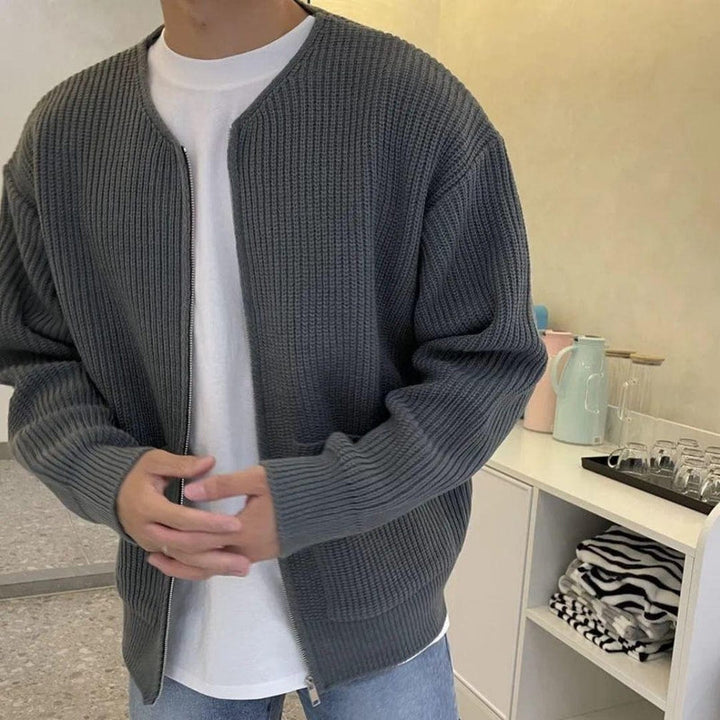 Uomo con cardigan grigio a maglia, t-shirt bianca e jeans, in cucina moderna con bollitore e bicchieri sullo sfondo. Moda casual maschile.
