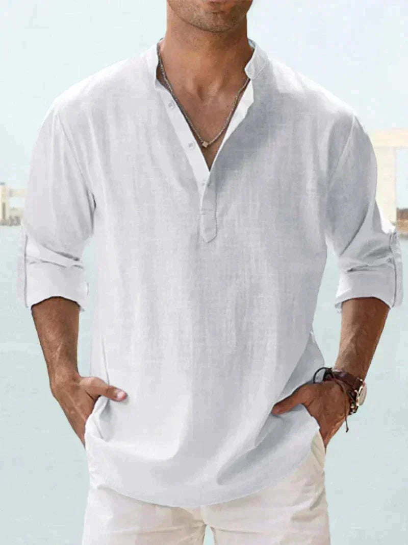 Camicia uomo bianca in lino, maniche lunghe arrotolate, collo alla coreana, stile casual estivo. Sfondo sfocato, look elegante e rilassato.
