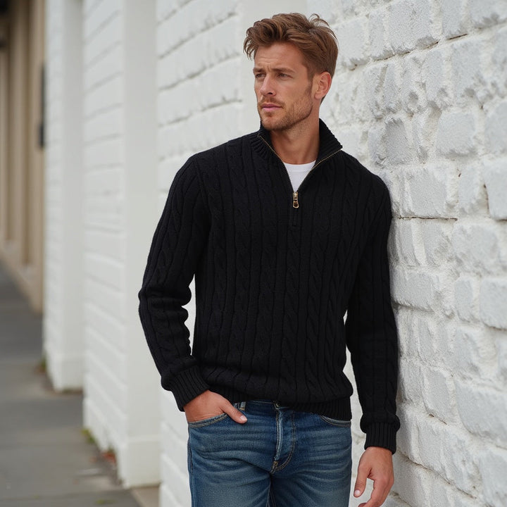 Uomo con maglione nero a trecce e jeans, appoggiato a un muro bianco. Moda maschile autunnale, stile casual elegante.