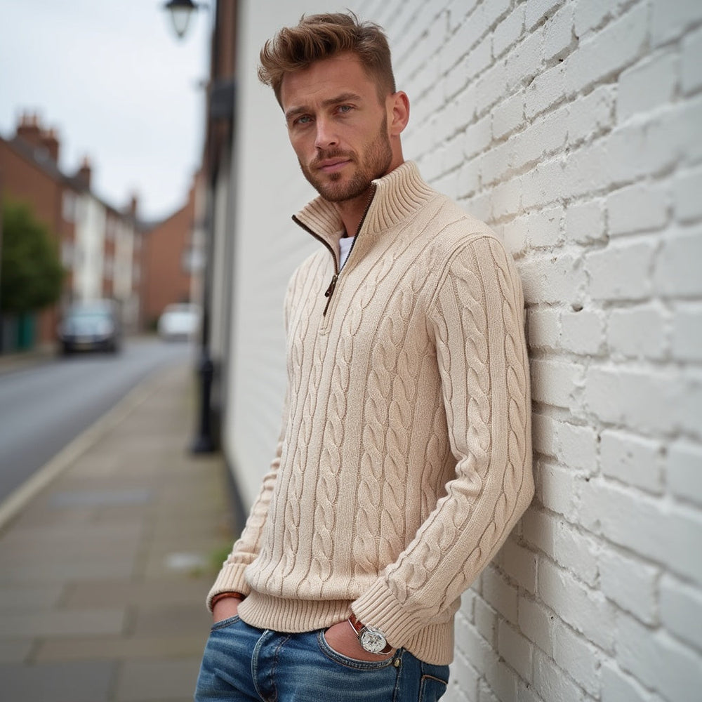 Uomo con maglione a trecce beige, appoggiato a un muro di mattoni bianchi, strada urbana sullo sfondo. Moda autunnale, stile casual elegante.